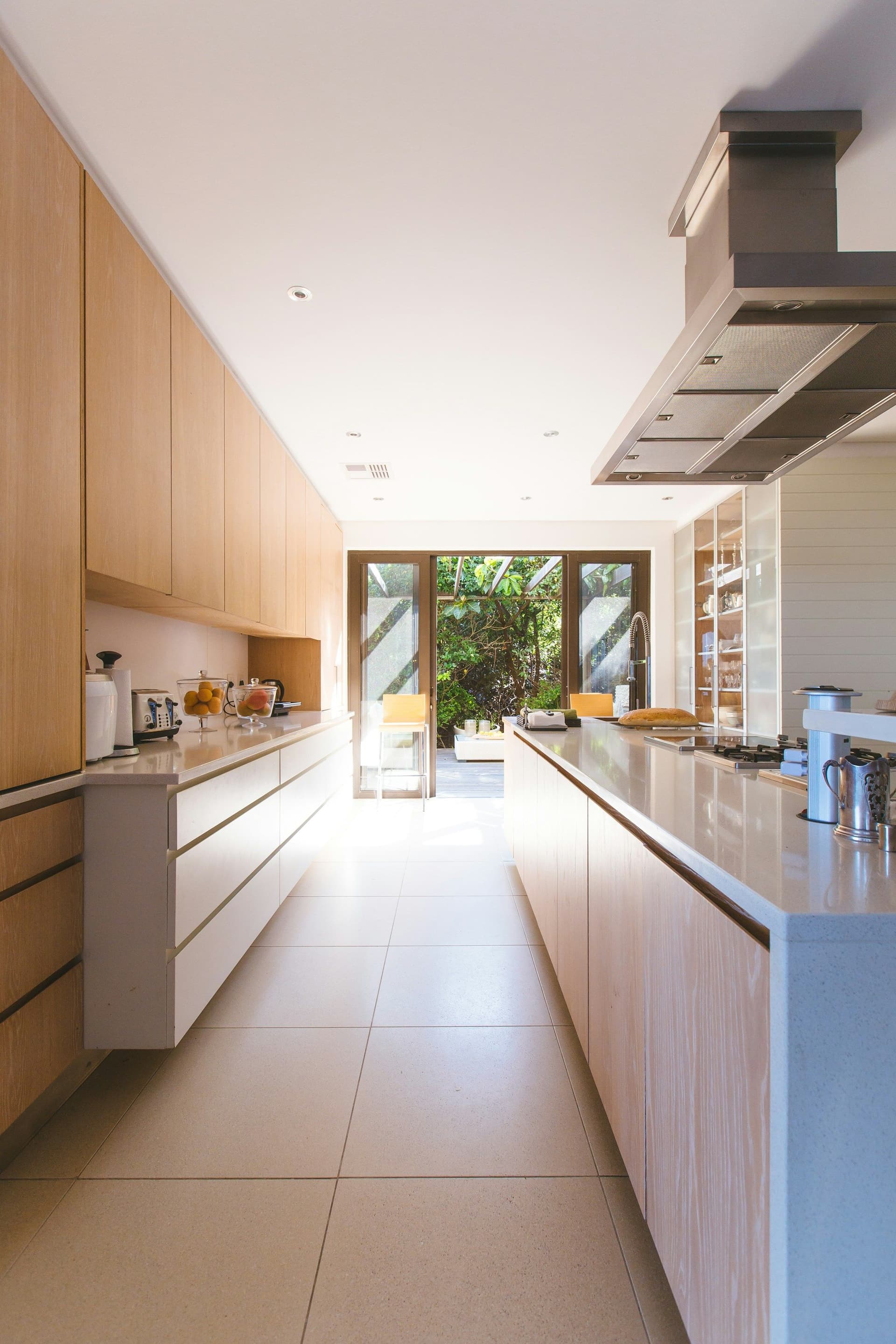 A long galley kitchen with light wood cabinets, a continuous countertop and glass doors opening at the far end