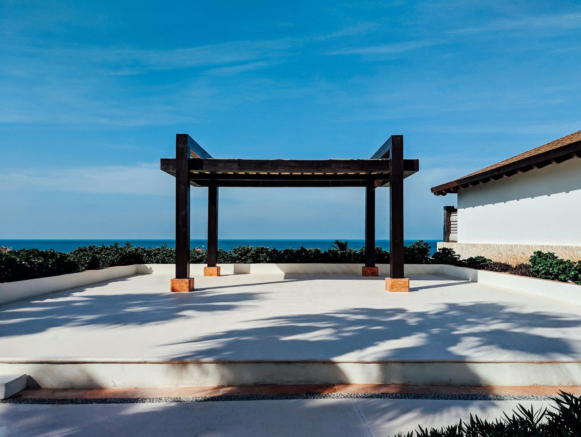 Black steel pergola frame on a pale concrete platform beside the sea under a blue sky