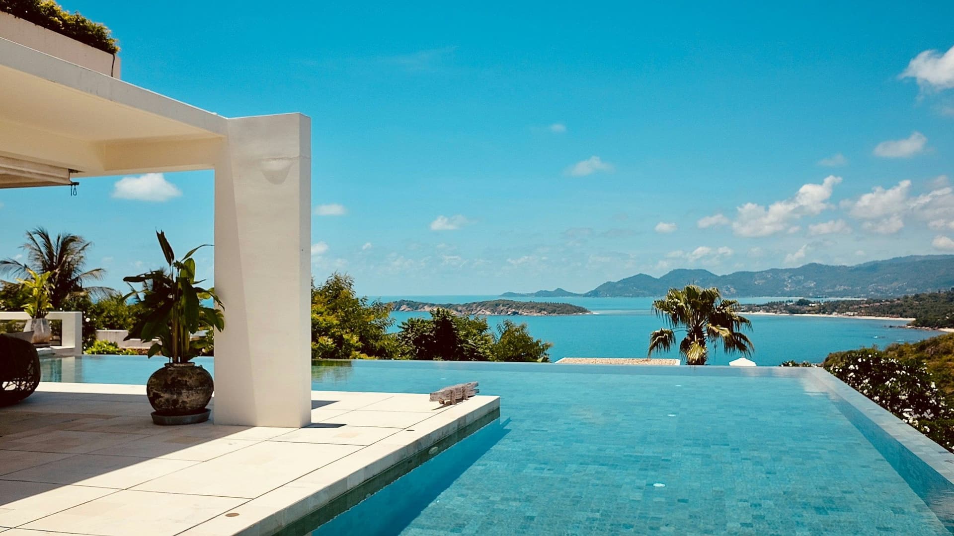 Turquoise infinity pool beside a white terrace, with a sea bay and islands beyond