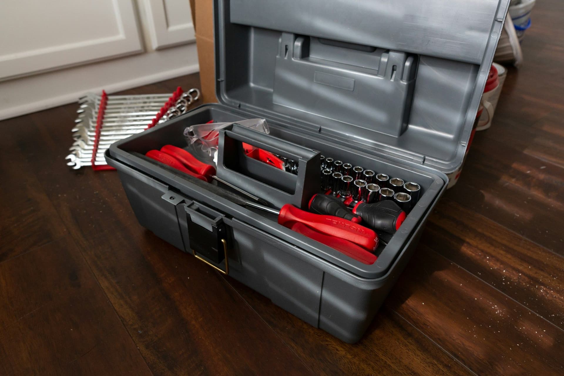An open gray toolbox with socket sets, pliers and red-handled tools on a dark wooden floor