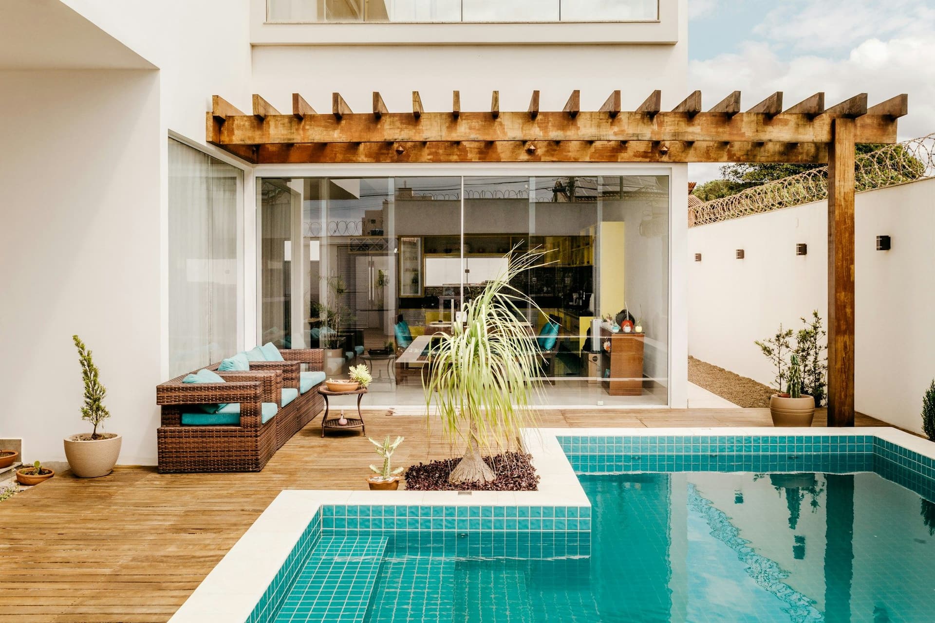 Wood-beam pergola over a wooden deck with wicker sofas and a teal-tiled pool beside a white house