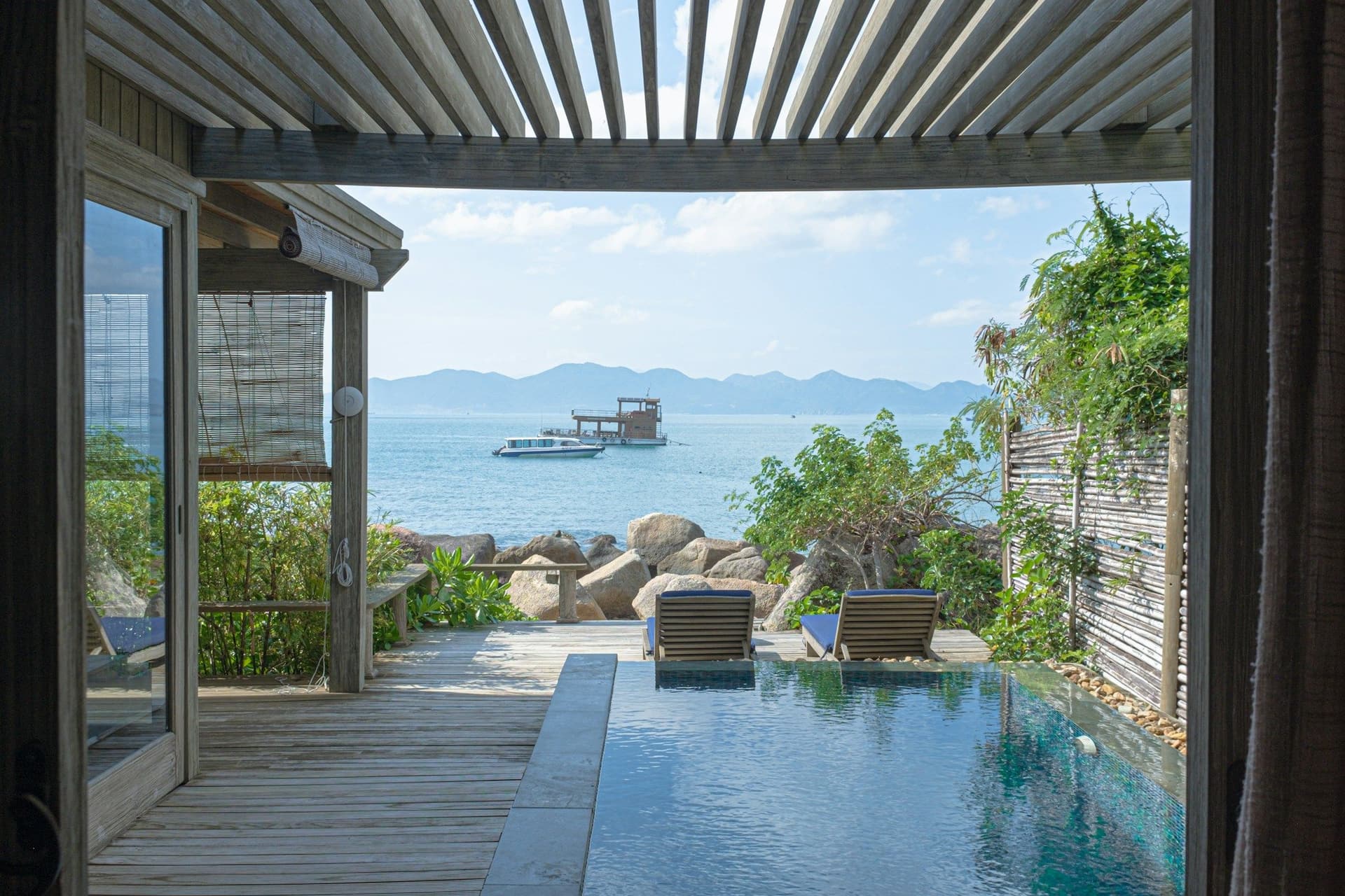Louvered metal pergola roof framing a wooden walkway, a narrow pool and a sea view