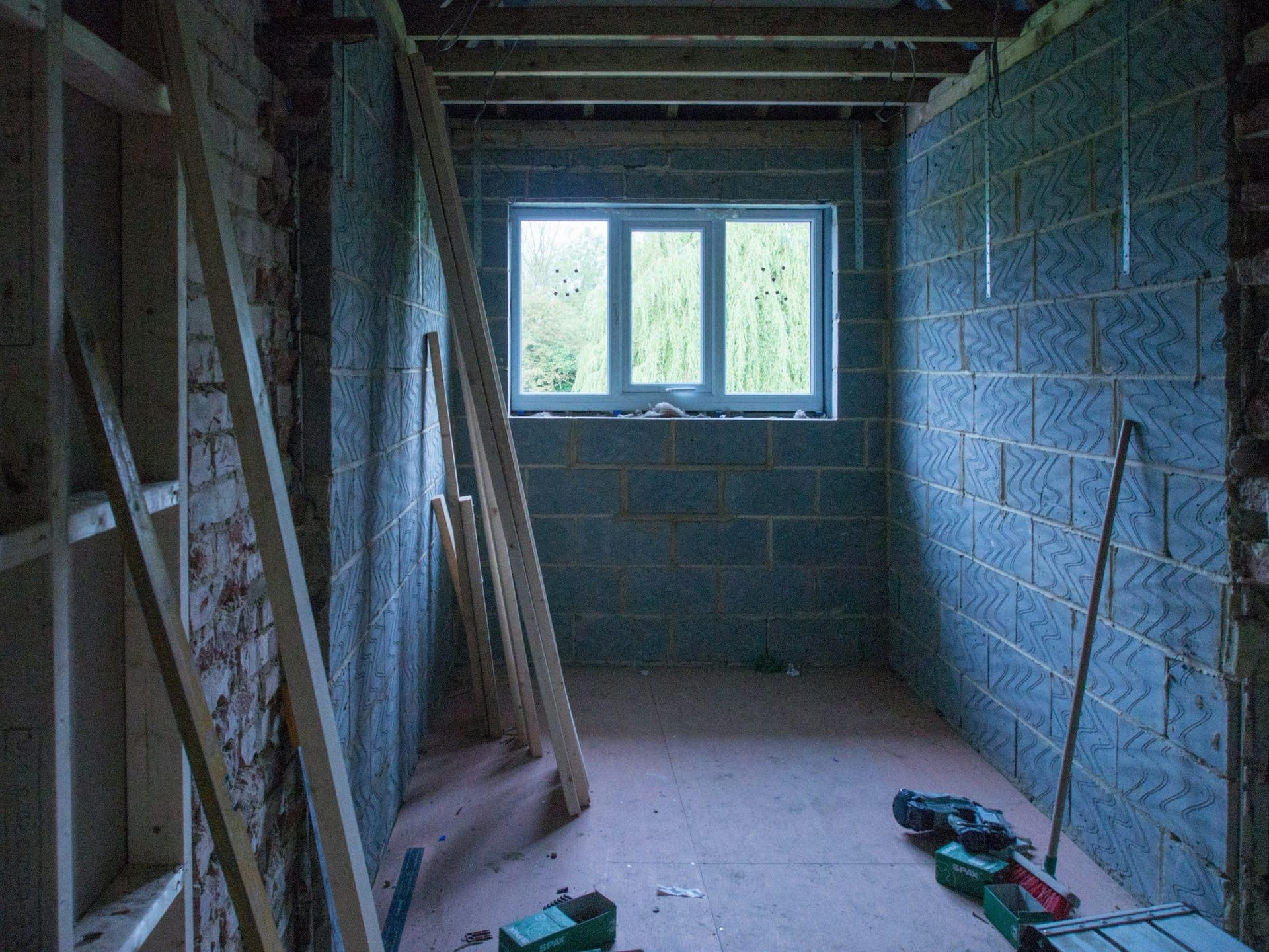 A room built in concrete block with a window, timber planks against the wall and a drill on the floor