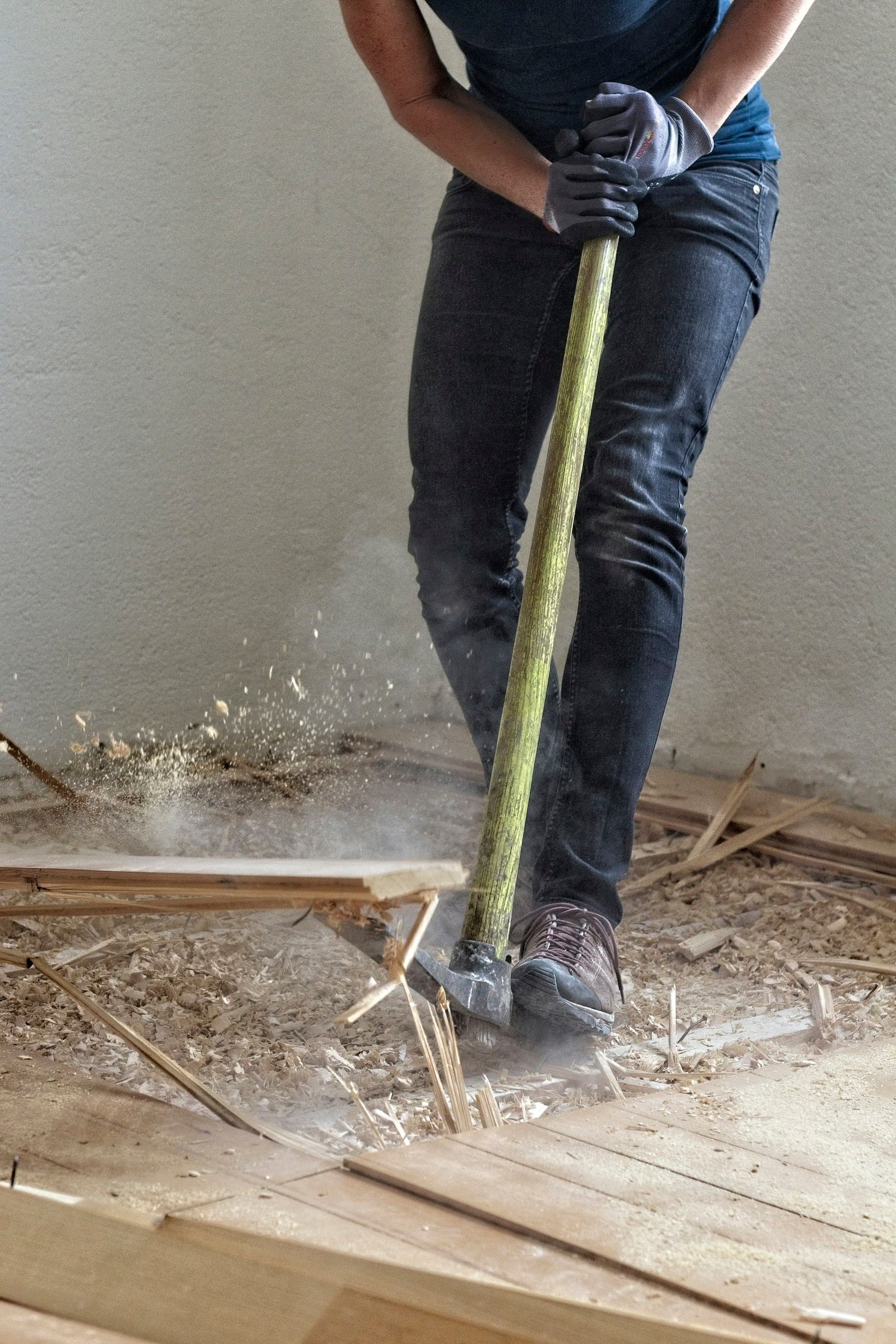 A person in work gloves swinging a sledgehammer at a wooden floor, with dust and splinters flying