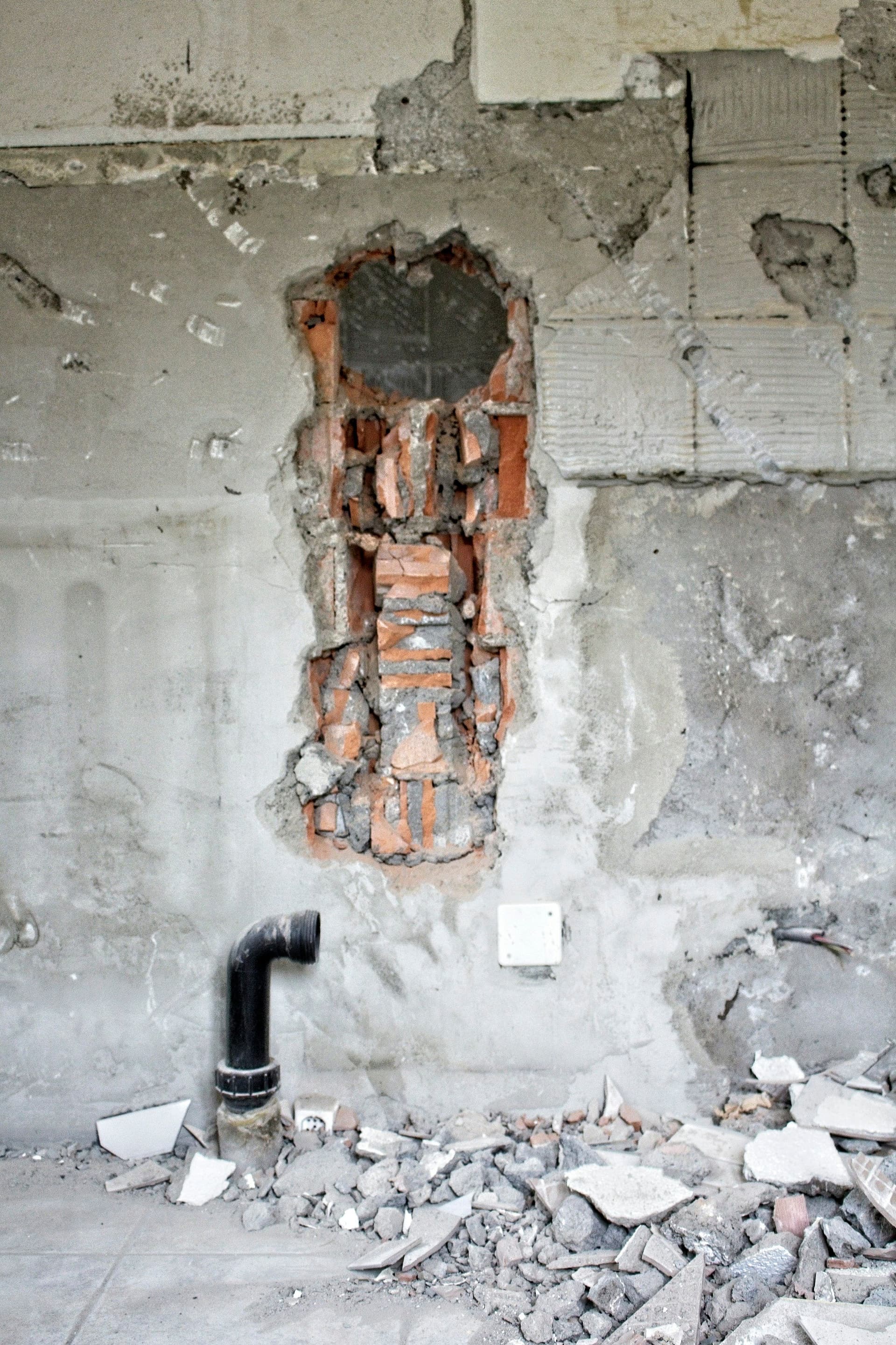 A plastered wall opened to show red brick, with a black pipe stub and rubble on the floor