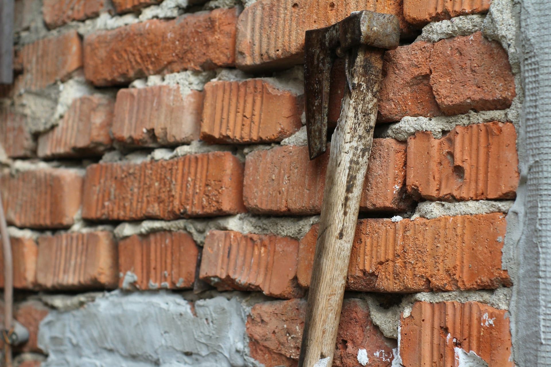 A hammer leaning against an exposed red brick wall with patches of gray mortar