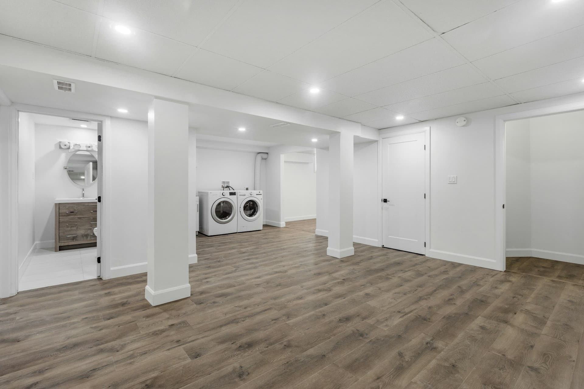 Finished basement room with white walls, recessed lights, support columns, a wood-look floor and a laundry area at the back