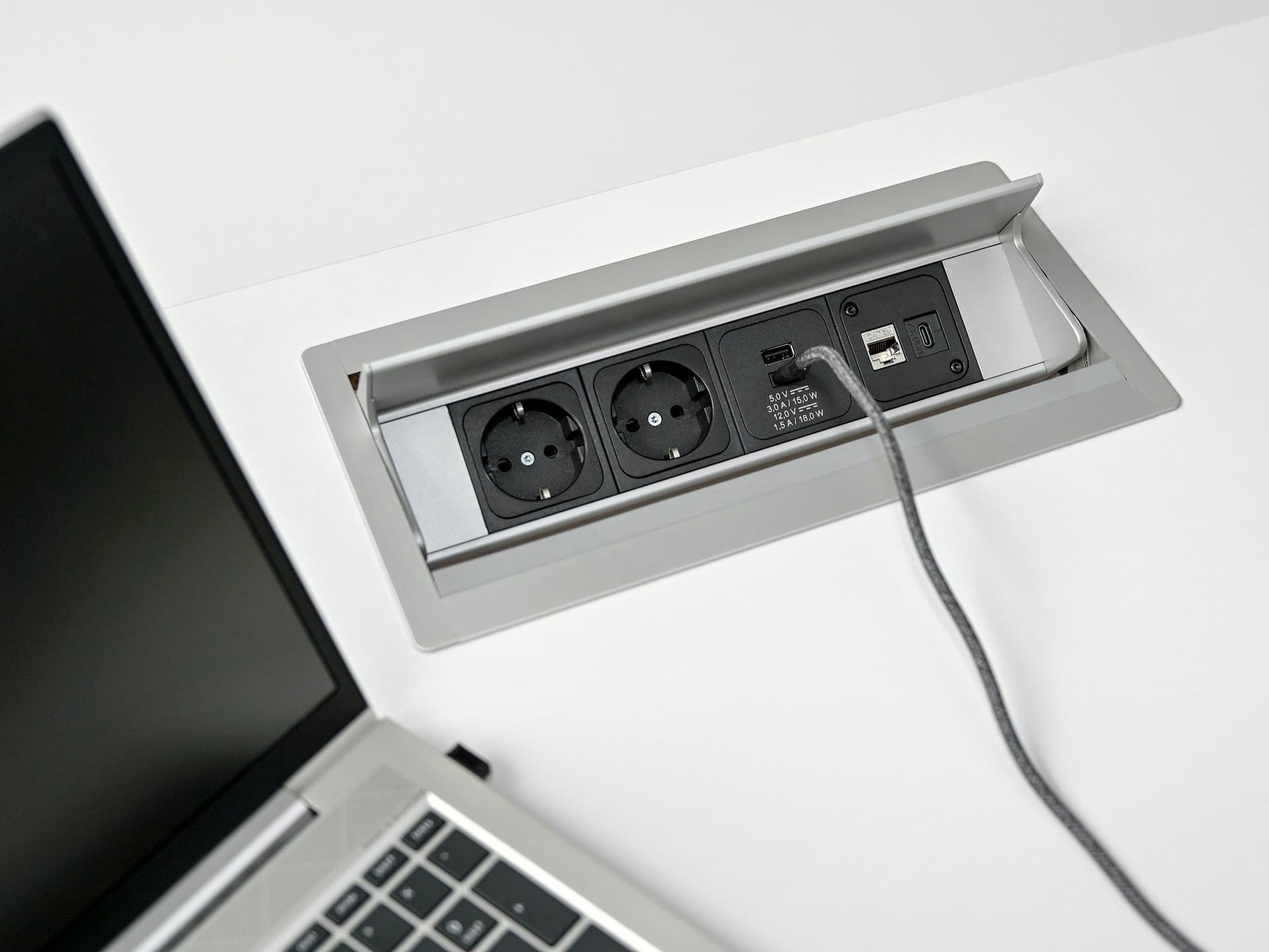 Power box set into a white desk, open to show two sockets and USB ports, next to a laptop