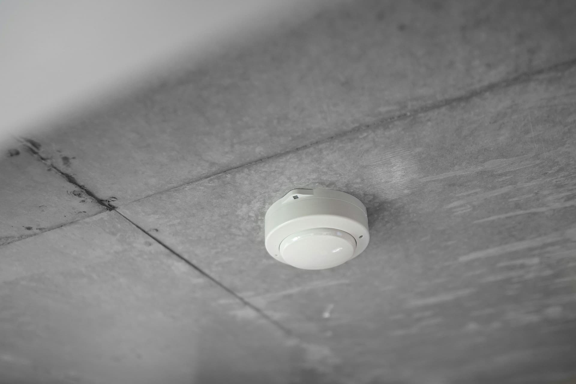 A small round white smoke detector mounted on a gray concrete ceiling in a black and white photo