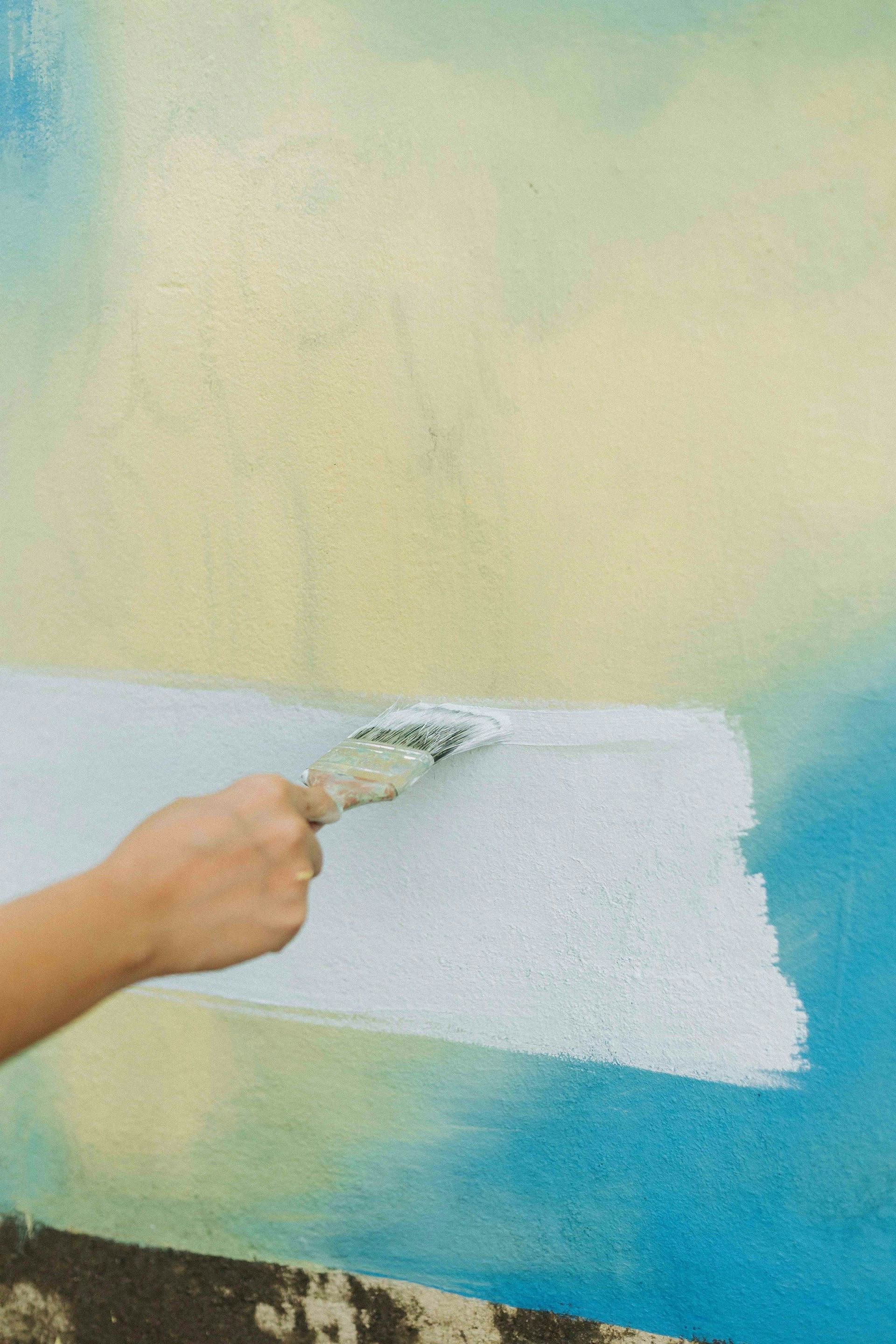 A hand brushing white paint over a wall that was previously painted in yellow and blue