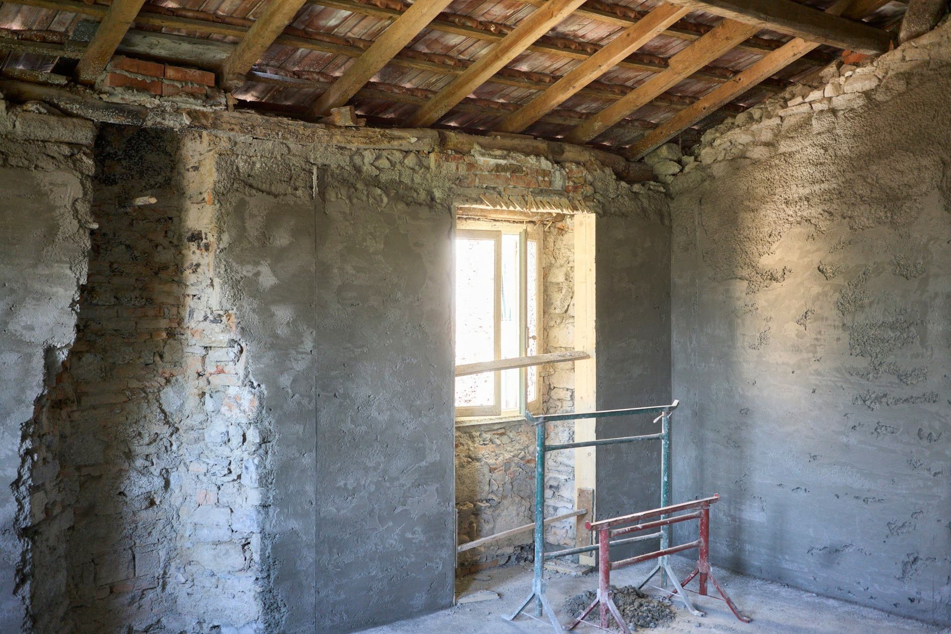 A bare room with rough plastered walls, exposed rafters overhead, a small window and two trestles on the floor