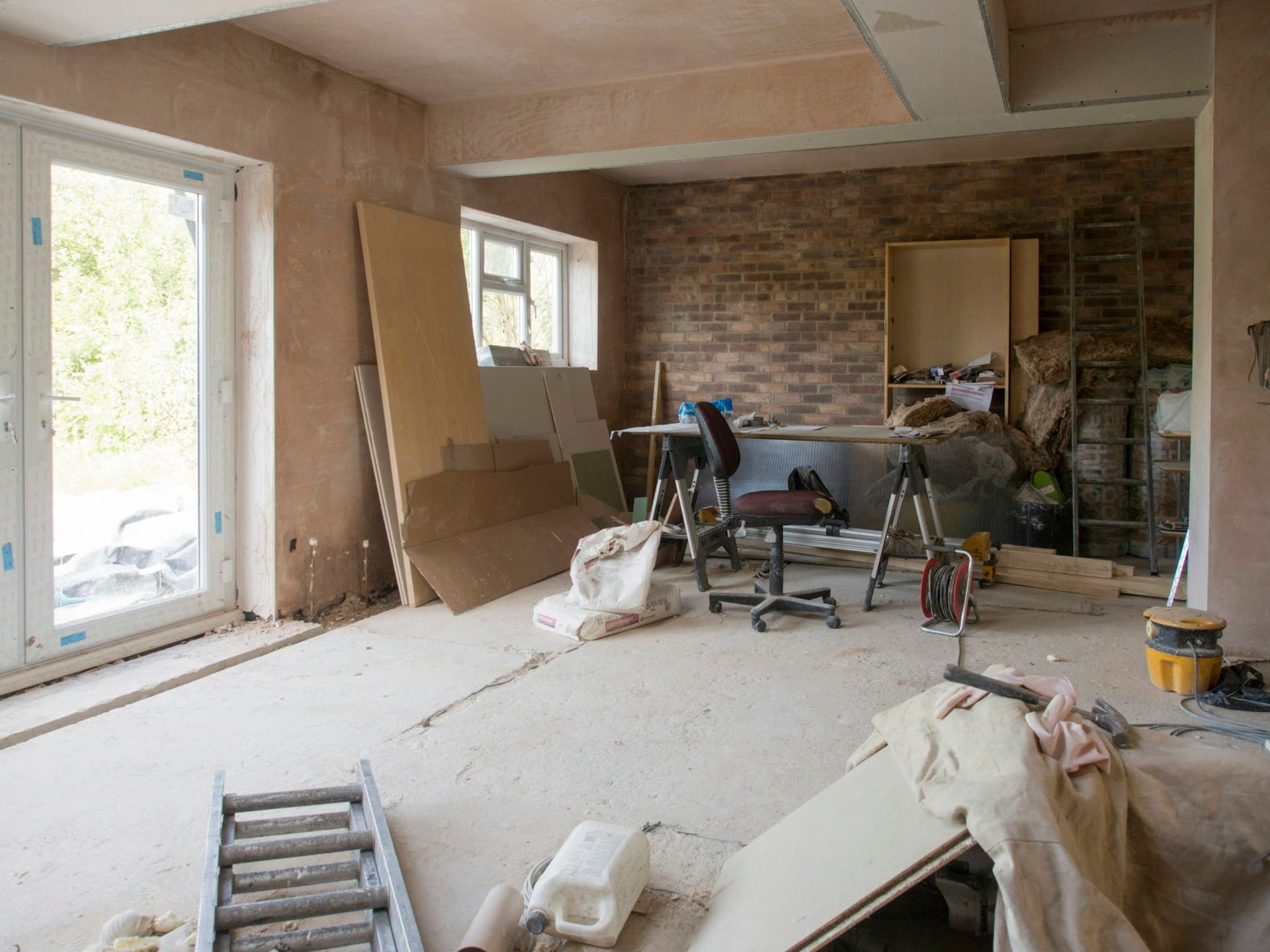 A room mid-renovation with a new glass door, plastered walls, boards leaning on the wall, a ladder and trestle tables
