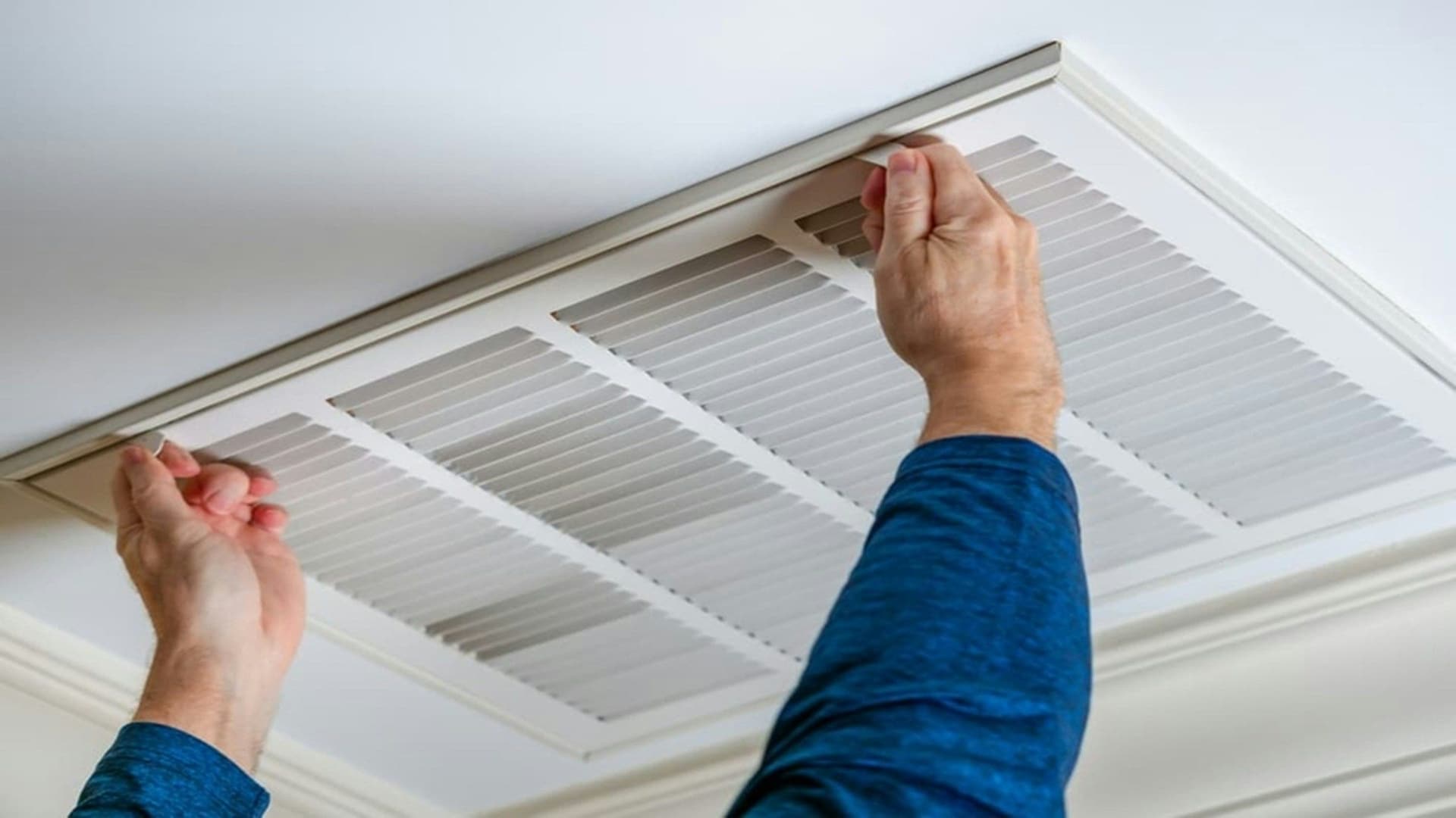 Two hands lifting a white ceiling air conditioning grille to reach the filter