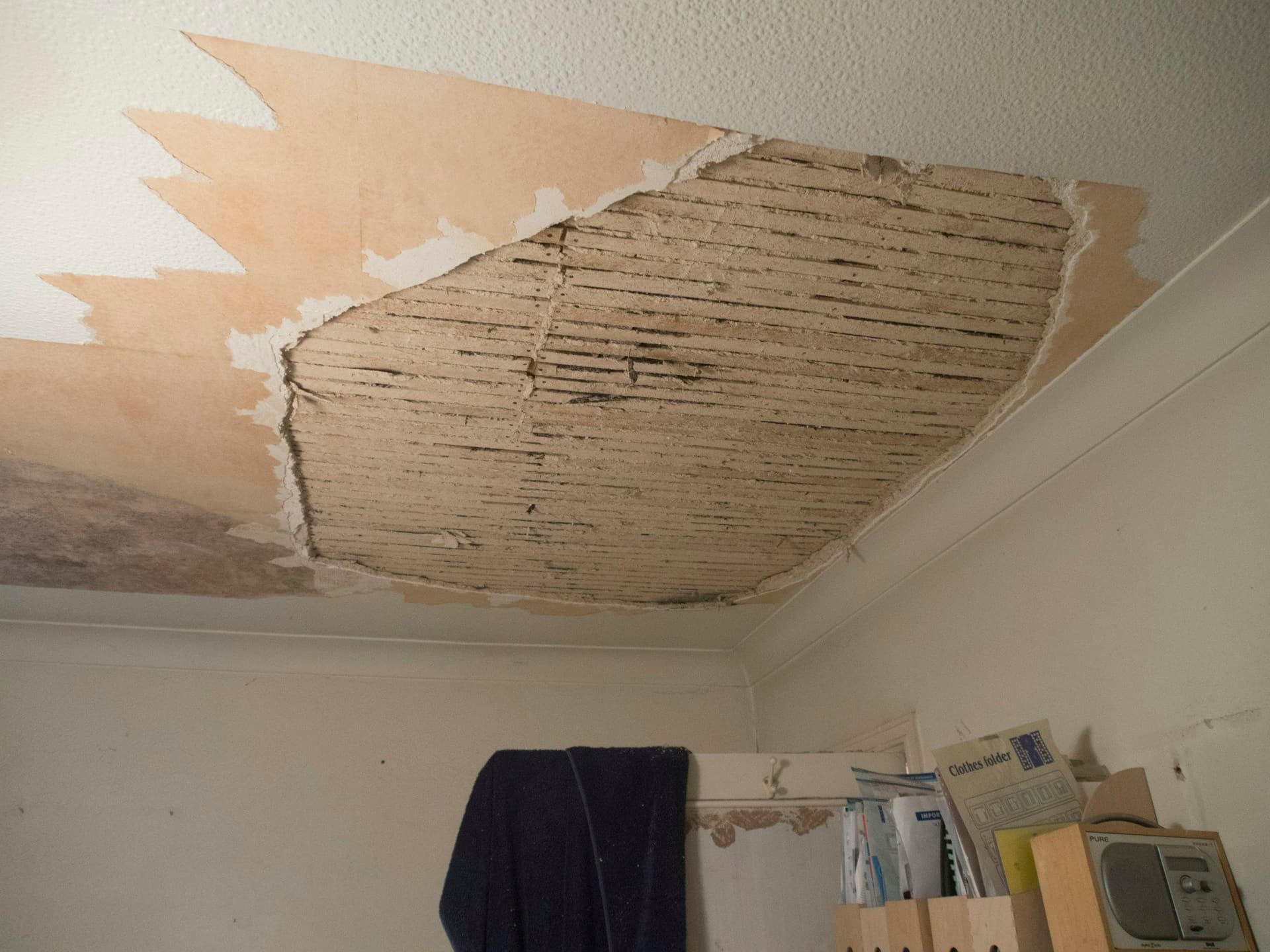 Room ceiling where a large patch of plaster and paper has peeled away, exposing the wooden laths above