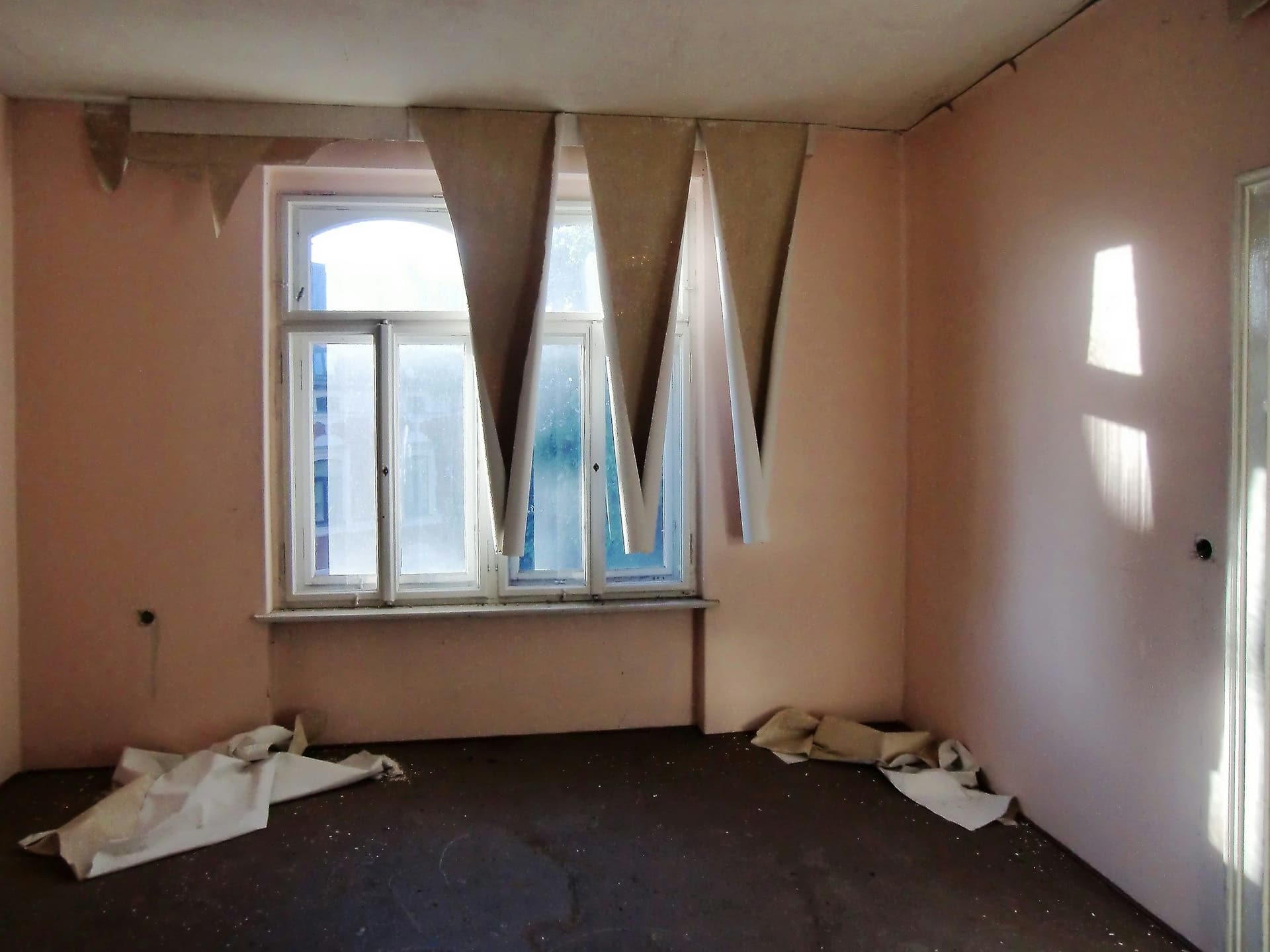 An empty room with pink walls, strips of old wallpaper hanging by the window and sheets on the floor