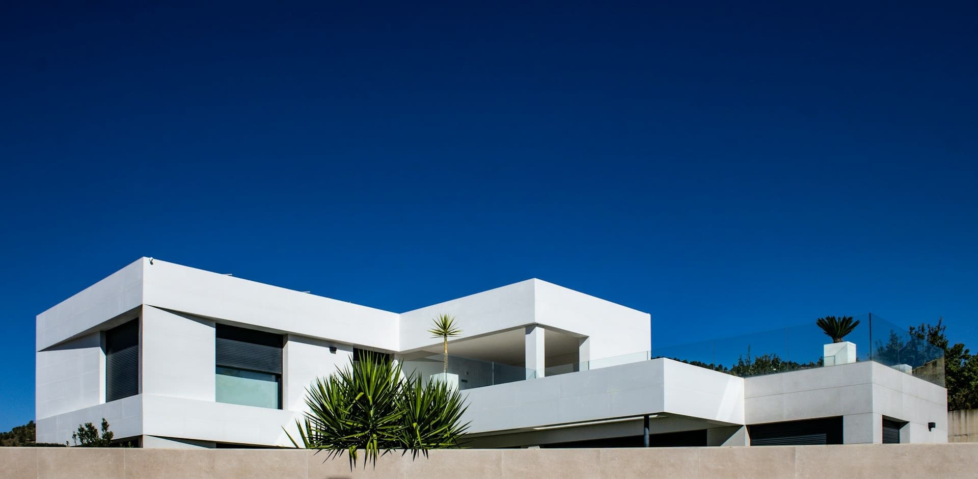 White modern villa with flat roofs and roof-level planting, seen from below against a deep blue sky