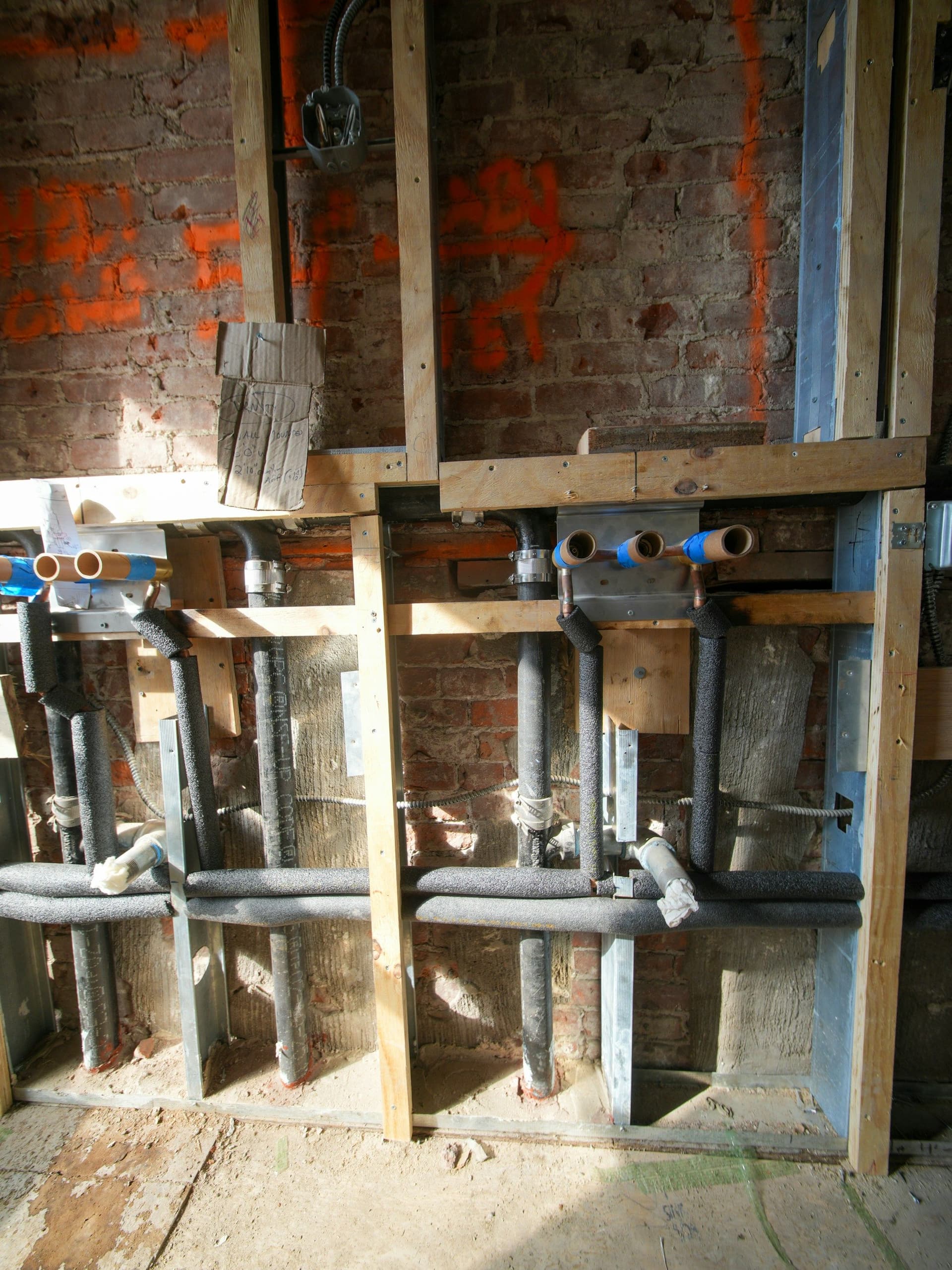 Exposed plumbing pipes and electrical conduit between wooden studs against a brick wall in a room under renovation