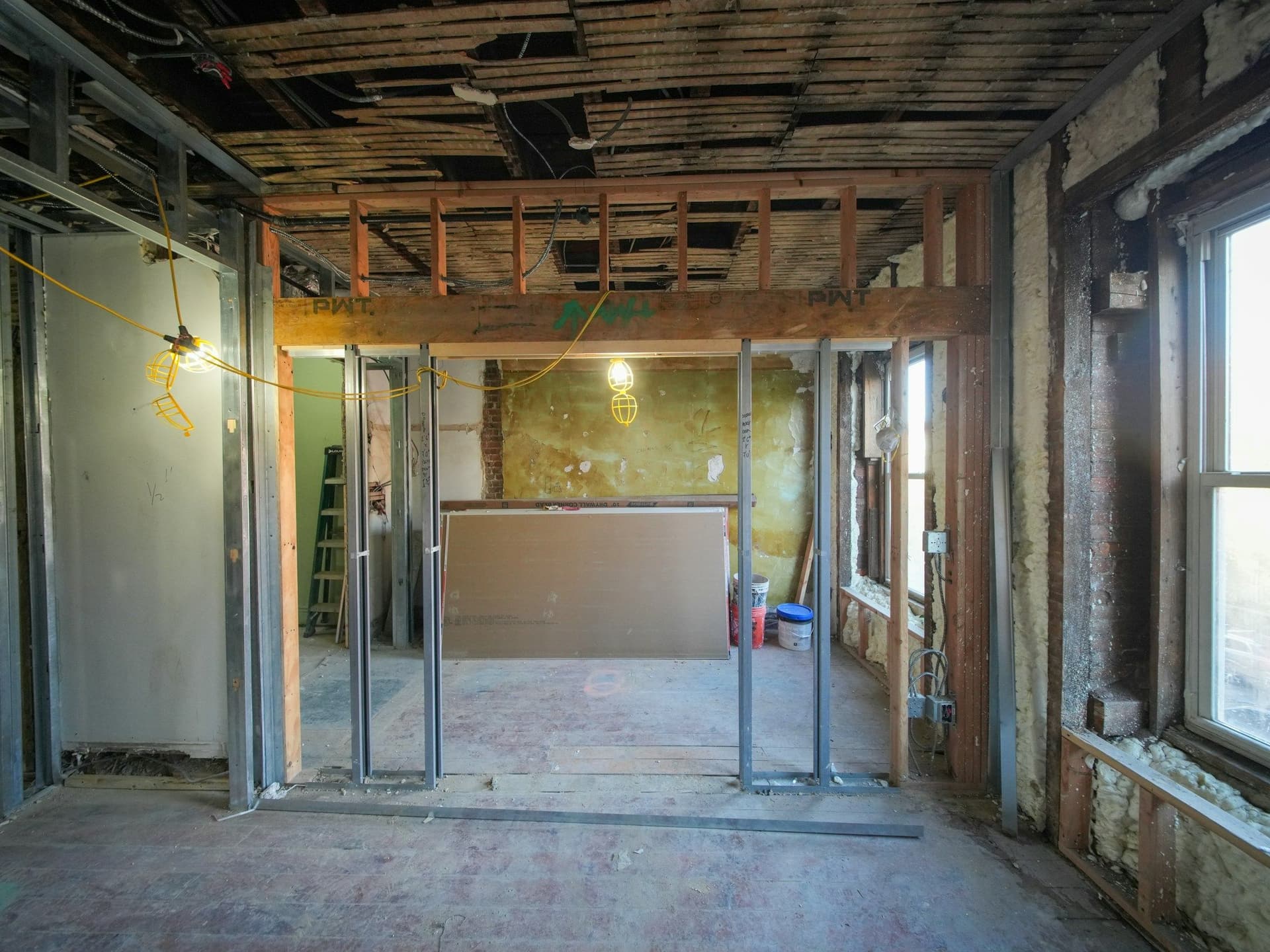 A room mid-renovation with metal wall studs, a timber beam, bare ceiling joists and a sunlit window
