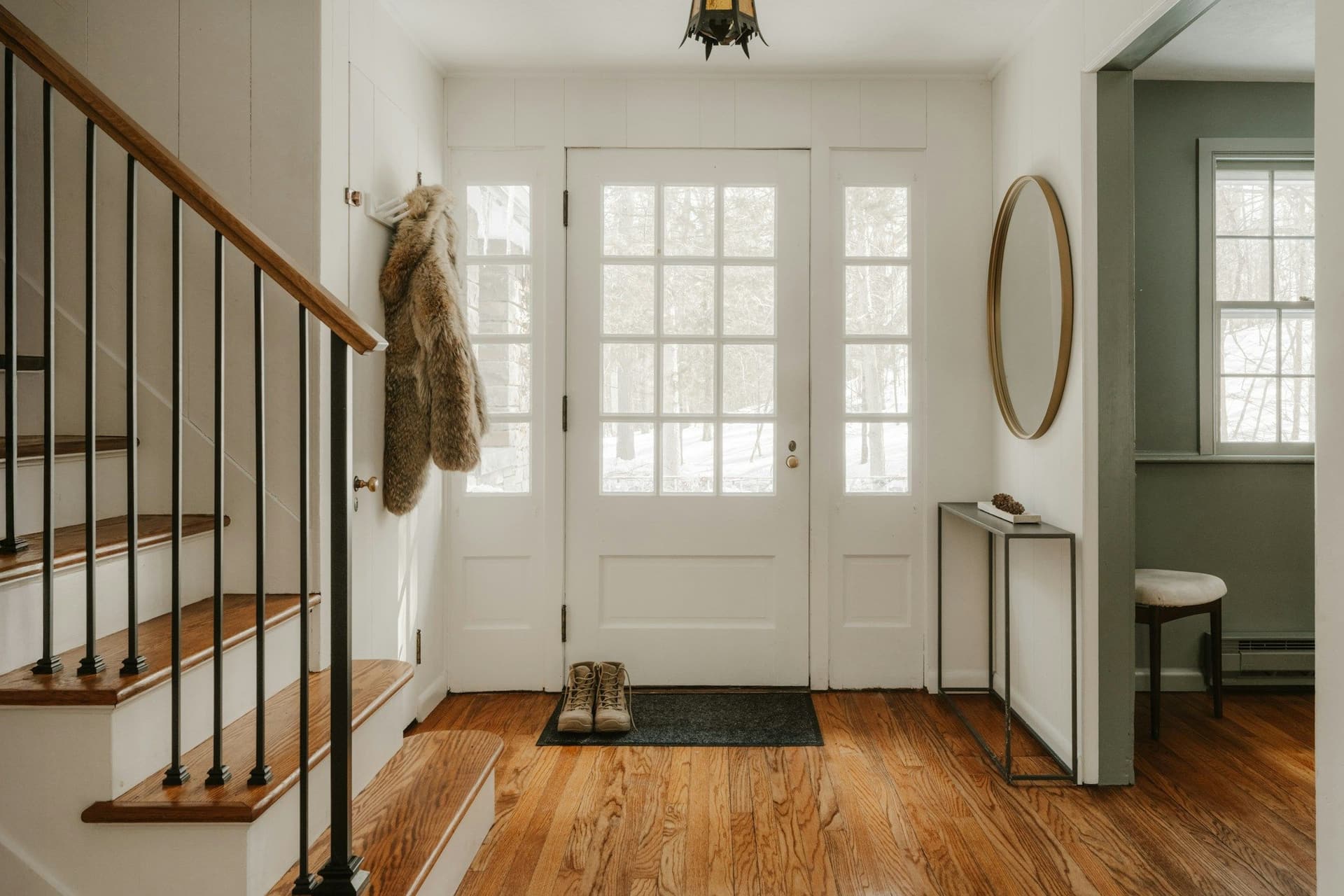 Entry hall with wooden stairs and black balusters beside a white glass-paneled front door and a round mirror