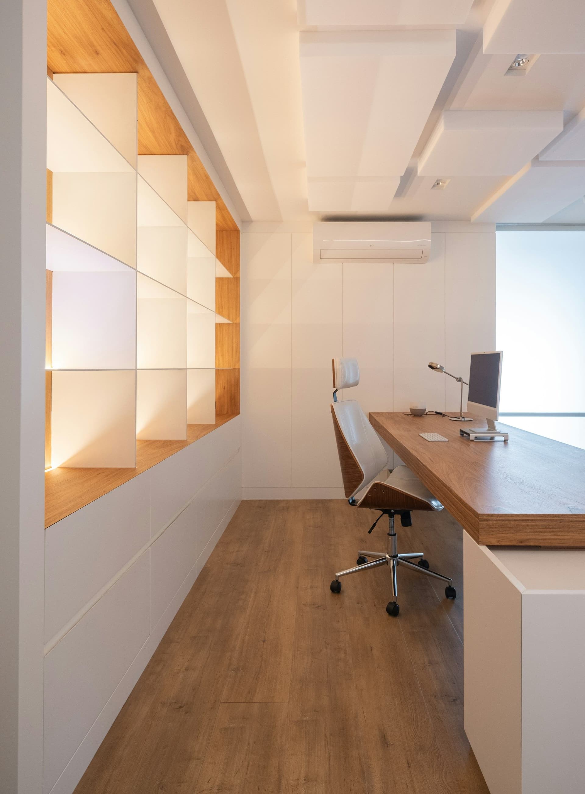 Long narrow office with a wooden desk, backlit white shelving, a wall-mounted air conditioner and a wood floor