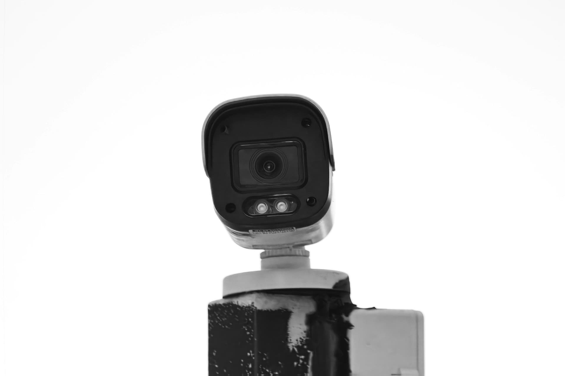White bullet-style security camera mounted on a dark post against a plain white sky