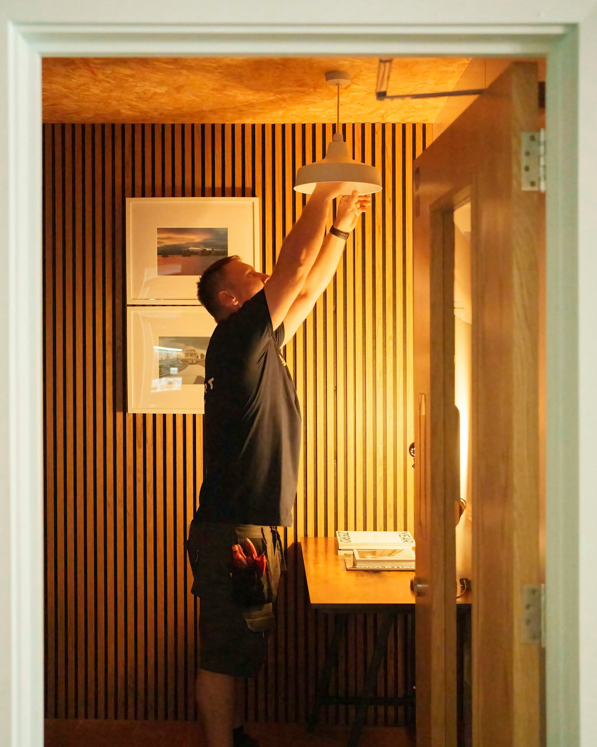 A man in a black T-shirt reaches up to fit a pendant light in a warm, wood-slatted room