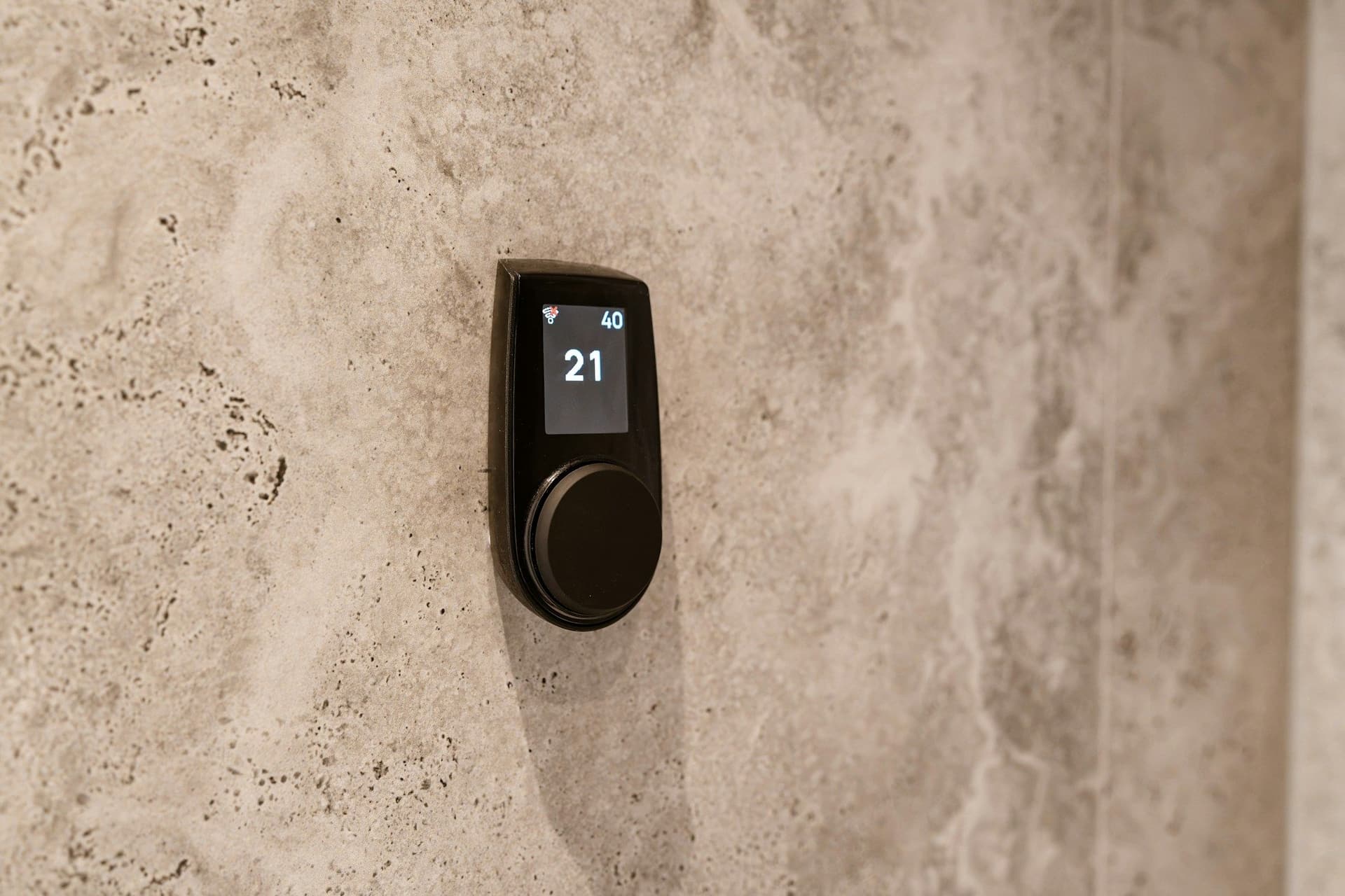 Slim black wall control with a temperature display and round dial on a pale stone-effect wall